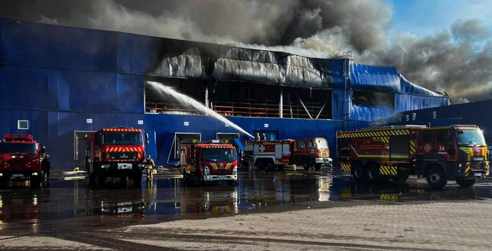 Fire engines and emergency crews spray water into a smoke-filled warehouse damaged near Kyiv