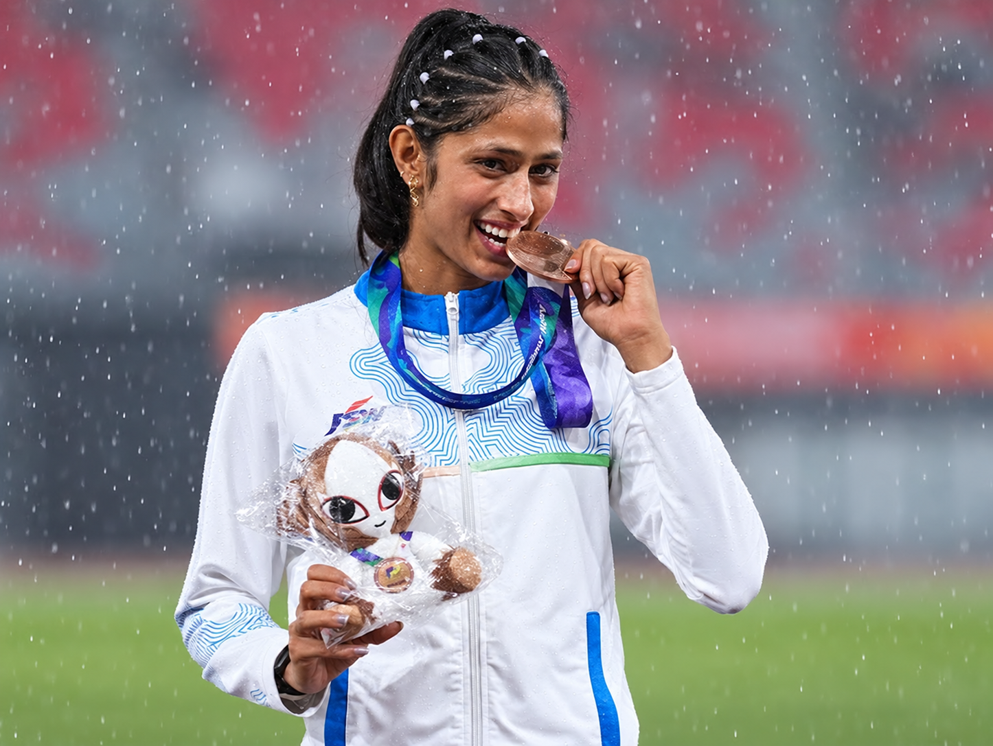 Pooja Singh bites her bronze medal while holding the Asian Games mascot in the rain.