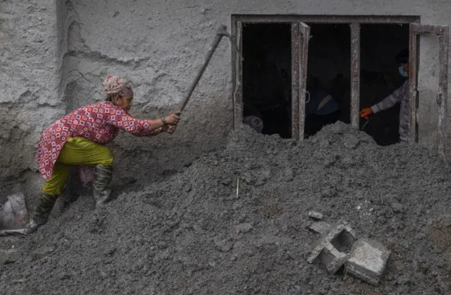 A woman clears thick flood mud piled against a building in Devighat, Nuwakot